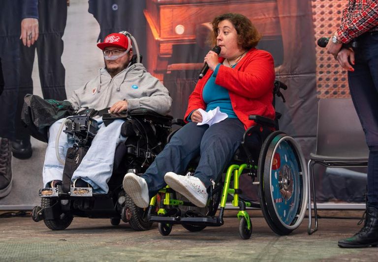 Zwei Personen mit Rollstühlen berichten von ihren Erfahrungen an der Behindertensession auf einer Bühne